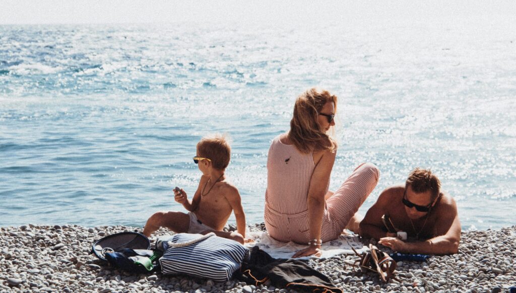 Vater, Mutter und ihr junger Sohn sitzen am Kiesstrand eines Sees.