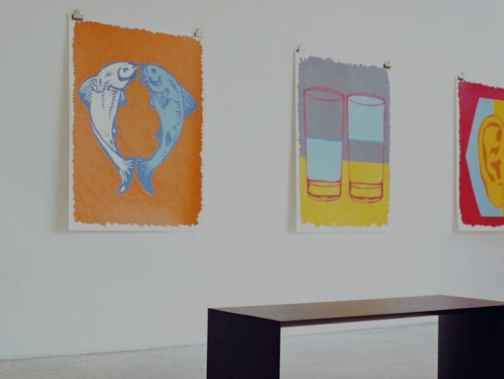 Moderne Malereien–ein orangenes Bild mit Fischen, zwei symmetrische Gläser–hängen an einer weißen Wand vor einer Bank.