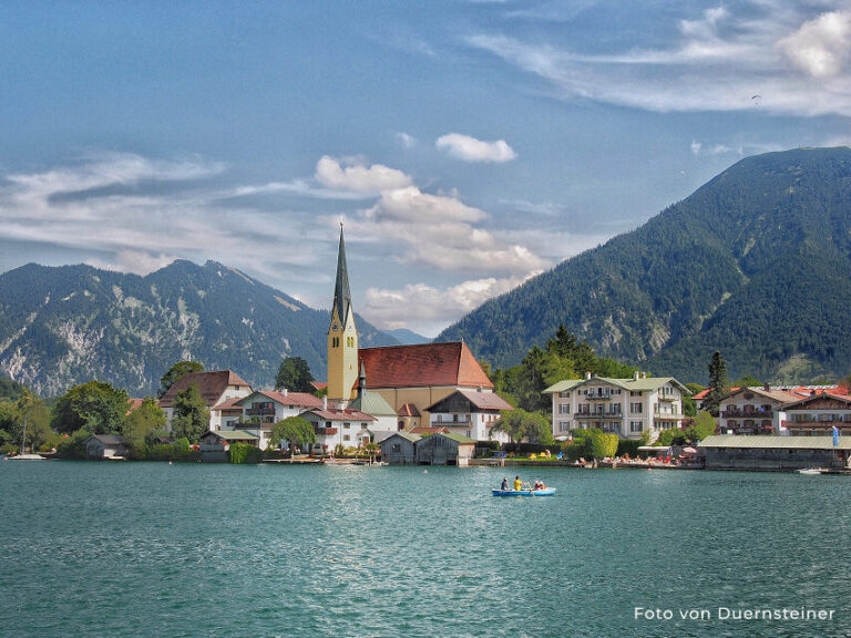 Bild von Tegernsee. Ein Bild von einer Kirche am See mit Bergen im Hintergrund