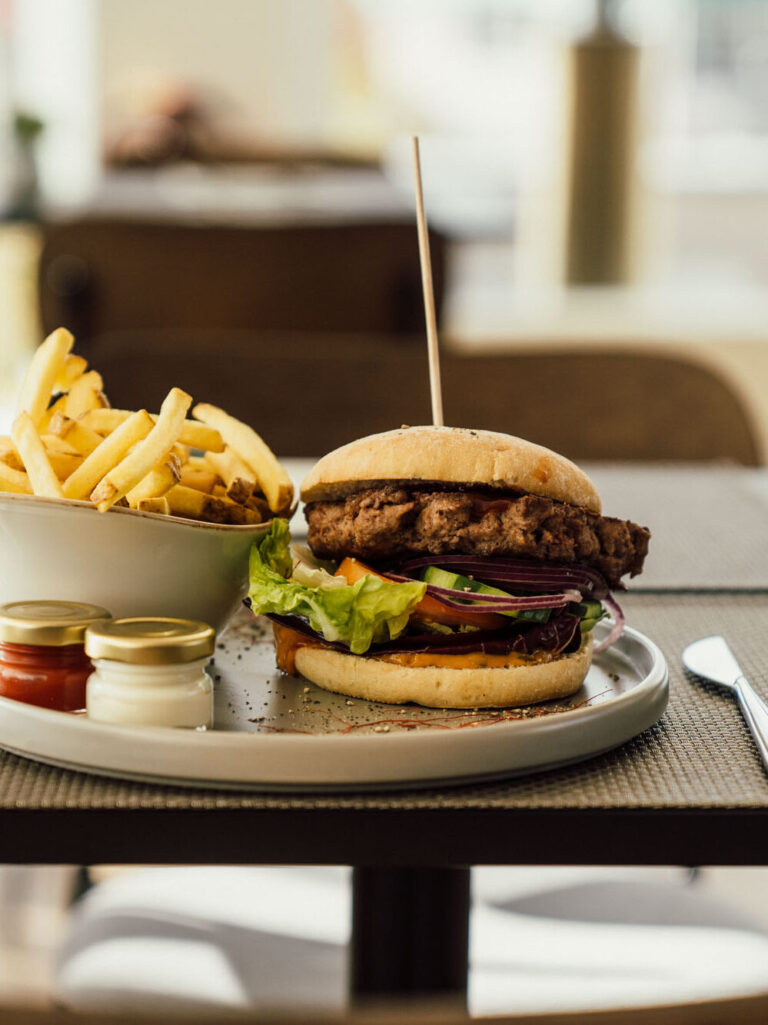 Darstellung eines Resturanttisches mit einem Teller auf dem ein Burger und dazugehörigen Pommes angerichtet sind.